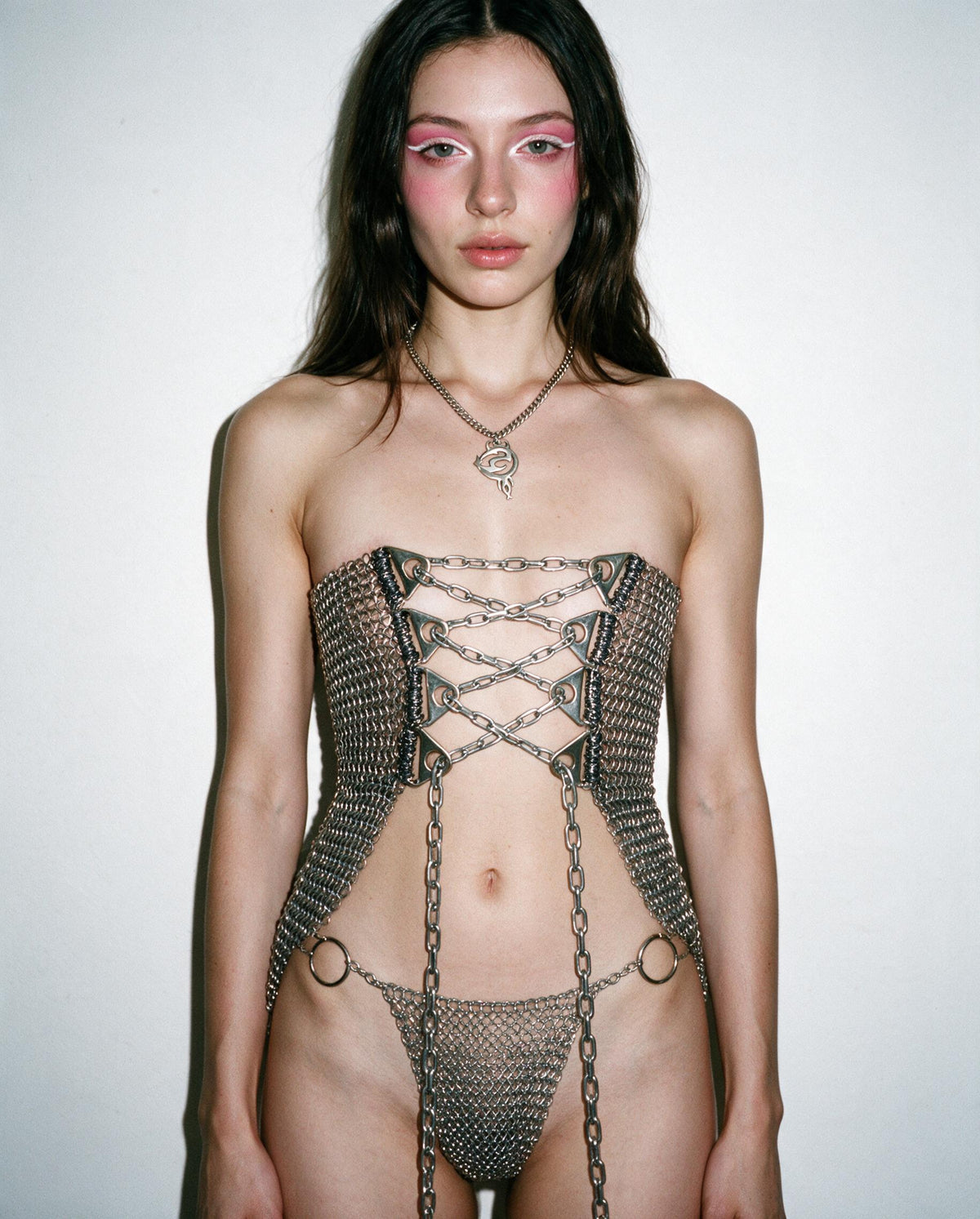 A model wearing a body-hugging chainmail corset with chain laces and matching chainmail thong, styled with a silver pendant necklace against a plain off-white background.