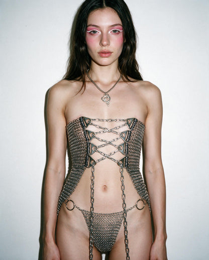 A model wearing a body-hugging chainmail corset with chain laces and matching chainmail thong, styled with a silver pendant necklace against a plain off-white background.